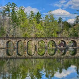 Stone arch bridge spanning a calm lake, with seven evenly spaced arches reflected almost perfectly in the still water. Tall pine and deciduous trees line the far shore beneath a bright blue sky with scattered clouds, creating a peaceful, mirror-like scene.