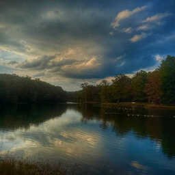 A calm lake reflects towering storm clouds stretching across the evening sky. Dense trees line both shores, their silhouettes framing the water as soft light breaks through gaps in the clouds, casting blue, gray, and golden tones across the landscape.