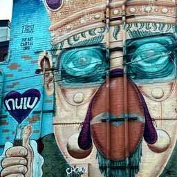 A colorful street mural transforms an entire building into the face of a crowned figure with oversized blue-green glasses, a long nose, and purple accents. Vertical drainpipes bisect the face, cleverly incorporated into the composition. A large hand on the left holds a purple heart emblazoned with "NULU," while "The Art Cartel" logo is painted near the top.