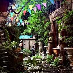 A lush courtyard filled with tropical plants, weathered stone statues, and carved wooden architectural pieces. Colorful triangular bunting hangs overhead, while Buddha statues, classical sculptures, and antique garden ornaments line a narrow garden path beneath a canopy of greenery.