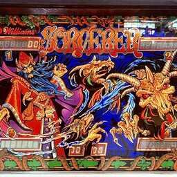 Backglass artwork from the 1985 Williams Sorcerer pinball machine. A blue-bearded wizard in flowing red and purple robes casts a spell while skeletal dragons and a horned dragon's skull erupt from swirling magical energy. Bright reds, blues, oranges, and purples create a dramatic fantasy scene around the game's score displays.