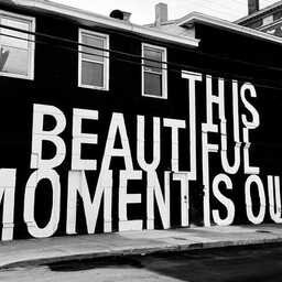 Black-and-white photograph of a building with a massive mural reading "THIS BEAUTIFUL MOMENT IS OURS" in oversized white block letters painted across the black exterior. Windows, doors, and the building's architecture are integrated into the typography.