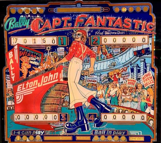 Colorful vintage Captain Fantastic pinball backglass featuring a flamboyantly dressed figure in a red star-patterned shirt, white bell-bottoms, oversized blue platform boots, knit cap, and round glasses, posing beside a bright red Elton John pinball machine. A cheering crowd, score displays, signs, stars, and carnival-like details fill the busy red, blue, turquoise, and orange artwork.