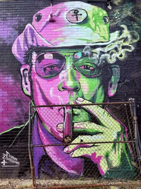 A surreal urban mural portrays a man wearing a hat, cigarette smoke drifting into the air as neon pink and green light washes across his face. The vibrant colors and oversized portrait dominate the dark wall behind an aging chain-link fence.