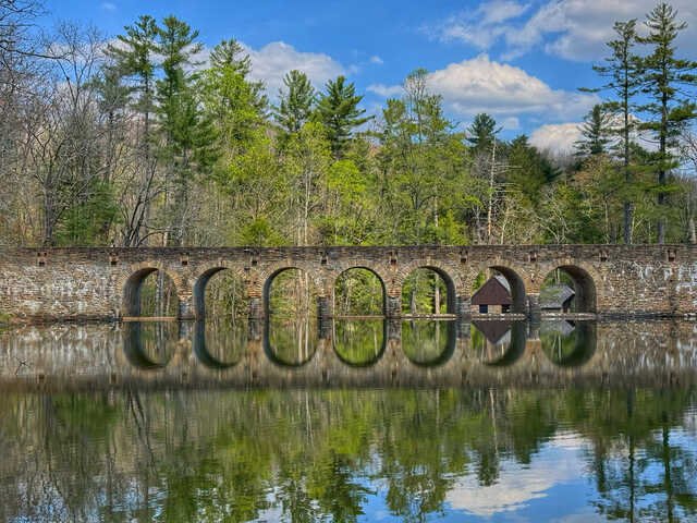 Stone arch bridge spanning a calm lake, with seven evenly spaced arches reflected almost perfectly in the still water. Tall pine and deciduous trees line the far shore beneath a bright blue sky with scattered clouds, creating a peaceful, mirror-like scene.