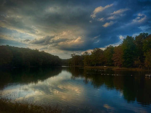 A calm lake reflects towering storm clouds stretching across the evening sky. Dense trees line both shores, their silhouettes framing the water as soft light breaks through gaps in the clouds, casting blue, gray, and golden tones across the landscape.