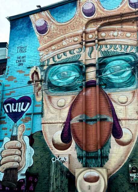 A colorful street mural transforms an entire building into the face of a crowned figure with oversized blue-green glasses, a long nose, and purple accents. Vertical drainpipes bisect the face, cleverly incorporated into the composition. A large hand on the left holds a purple heart emblazoned with "NULU," while "The Art Cartel" logo is painted near the top.