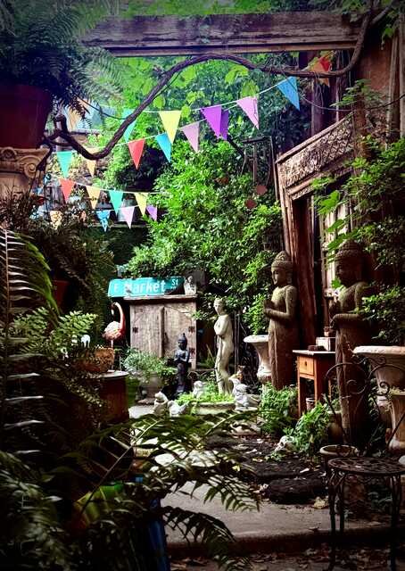 A lush courtyard filled with tropical plants, weathered stone statues, and carved wooden architectural pieces. Colorful triangular bunting hangs overhead, while Buddha statues, classical sculptures, and antique garden ornaments line a narrow garden path beneath a canopy of greenery.