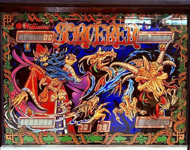 Backglass artwork from the 1985 Williams Sorcerer pinball machine. A blue-bearded wizard in flowing red and purple robes casts a spell while skeletal dragons and a horned dragon's skull erupt from swirling magical energy. Bright reds, blues, oranges, and purples create a dramatic fantasy scene around the game's score displays.