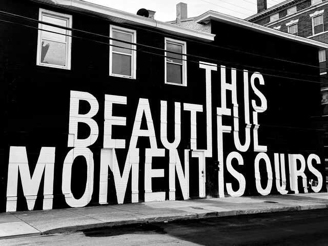Black-and-white photograph of a building with a massive mural reading "THIS BEAUTIFUL MOMENT IS OURS" in oversized white block letters painted across the black exterior. Windows, doors, and the building's architecture are integrated into the typography.